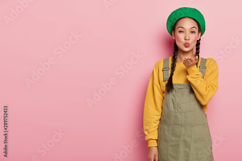Beautiful young female with Asian appearance, blows air kiss, keeps lips rounded, wears makeup, expresses love to someone, wears fashionable clothes, isolated on pink studio wall. Body language