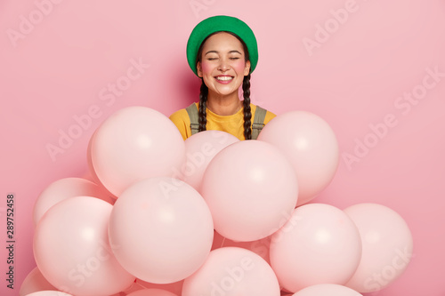 Portrait of happy Asian lady in green hat, has two braids, rouge cheeks, expresses positive emotions stands near many one colored balloons, celebrates Mums anniversary, isolated on pink studio wall