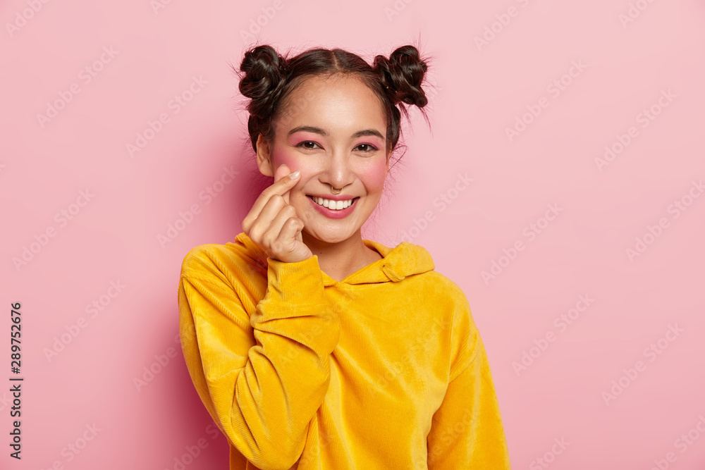 Portrait of happy Asian lady with pleasant smile on face, makes like sign, shapes heart with fingers, wears yellow sweatshirt, isolated over pink studio wall. Body language and valentine symbol Portrait of happy Asian lady with pleasant smile on face, makes like sign, shapes heart with fingers, wears yellow sweatshirt, isolated over pink studio wall. Body language and valentine symbol