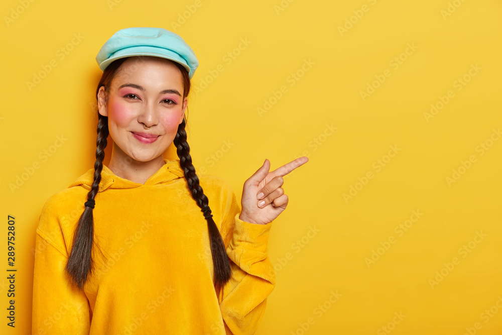 Look at copy space. Pleased cheerful Korean female model points aside, shows cool blank space, wears makeup, has two plaids, wears cap and yellow sweatshirt, poses indoor. Advertisement concept Look at copy space. Pleased cheerful Korean female model points aside, shows cool blank space, wears makeup, has two plaids, wears cap and yellow sweatshirt, poses indoor. Advertisement concept