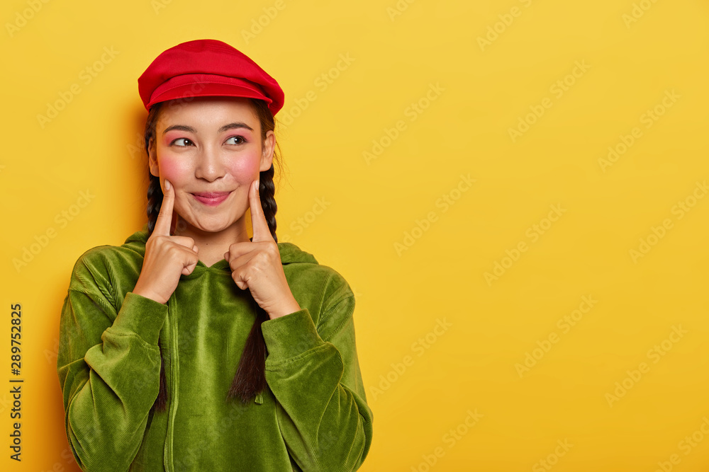 Pretty young Asian model keeps both fore fingers on cheeks, looks with dreamy expression aside, has minimal makeup, wears red beret, green velvet hoody, poses over yellow wall with blank space Pretty young Asian model keeps both fore fingers on cheeks, looks with dreamy expression aside, has minimal makeup, wears red beret, green velvet hoody, poses over yellow wall with blank space