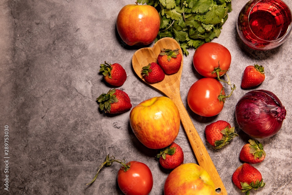 Fresh organic red themed fruit, vegetables, wine, and wooden heart shaped spoon, laying on concrete background countertop. Captured from above. Fresh organic red themed fruit, vegetables, wine, and wooden heart shaped spoon, laying on concrete background countertop. Captured from above.