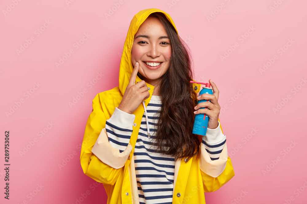 Happy brunette lady cures sore throat with spray, dressed in yellow raincoat with hoody, being ill after spending long time outdoor during rainy day, isolated on pink wall. Asian girl holds medicament Happy brunette lady cures sore throat with spray, dressed in yellow raincoat with hoody, being ill after spending long time outdoor during rainy day, isolated on pink wall. Asian girl holds medicament