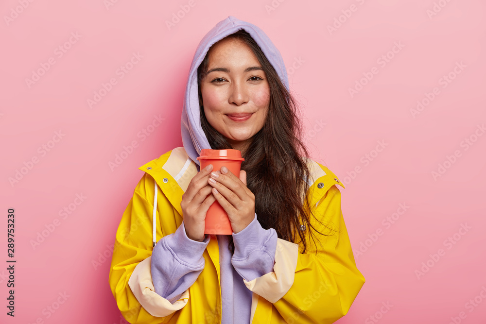 Pleased millennial girl with Asian appearance, has no makeup, wears violet sweatshirt and raincoat, holds flask with hot beverage, tries to warm while drinking tea, feels relaxed during rainy day Pleased millennial girl with Asian appearance, has no makeup, wears violet sweatshirt and raincoat, holds flask with hot beverage, tries to warm while drinking tea, feels relaxed during rainy day