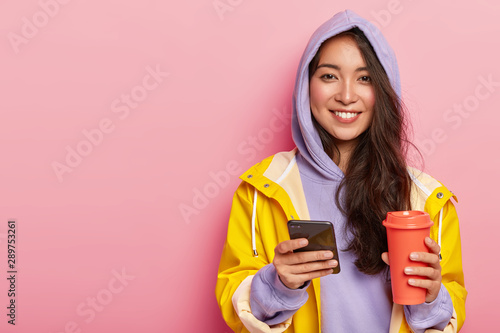 Delighted attractive Asian woman wears casual clothes, raincoat, sends message via cell phone, drinks aromatic beverage from thermos, being in good mood, always in touch. Season, leisure, lifestyle