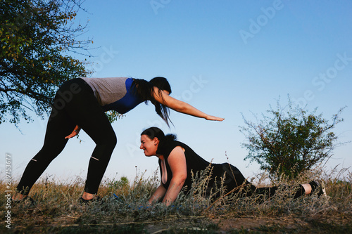 Personal trainer correcting her client at street workout. Overweight woman doing push-ups out with instructor support.