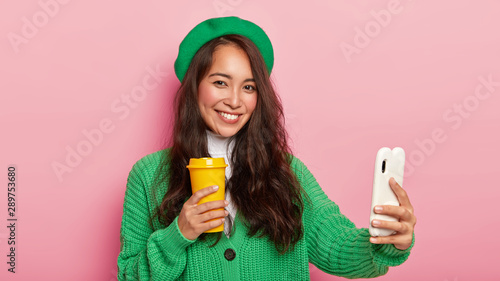 Photo of brunette Japanese woman makes selfie on modern smartphone, enjoys drinking hot beverage, dressed in bright vivid clothes, has tender appealing smile on face, poses indoor. Taking pics © wayhome.studio 