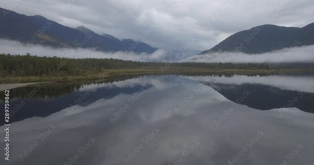 Fog Over Lake and Coastal Forest Under Southern Alps and Mirror Reflection on Water. Cinematic Aerial of New Zealand West Coast Fog Over Lake and Coastal Forest Under Southern Alps and Mirror Reflection on Water. Cinematic Aerial of New Zealand West Coast