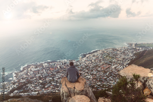 Young man sitting on the edge at the top of Lion's head mountain in Cape Town with a beautiful evening city view