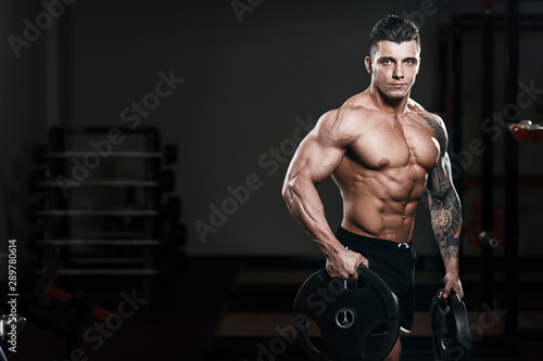 Muscular tattoo man bodybuilder training in gym and posing. Fit muscle guy workout with weights and barbell