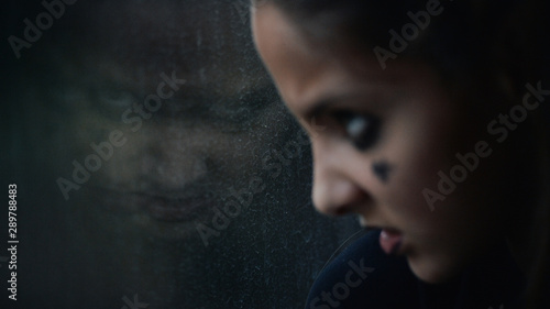 angry face of young little caucasian girl wearing dark youth subculture makeup watching at her sinister reflecton in dark window photo
