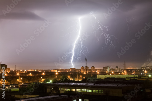 A powerful lightning strike hits the city at night. A strong lightning strike over a dark gray sky hits the ground, illuminating the industrial area around.