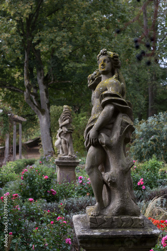 An old statue of a goddess stands in a botanical garden in Rothenburg ob der Tauber, covered with green moss, among the flowers.