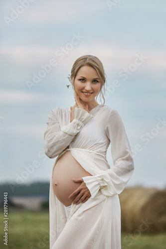 Pregnant girl walks in a field near haystacks in a long white dress, a woman smiles and holds her hands over her stomach. Girl is expecting the birth of a baby in the ninth month of pregnancy