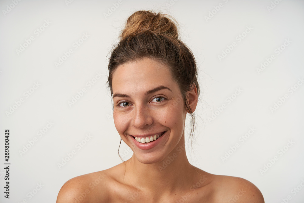 Indoor portrait of joyful young female smiling sincerely to camera, looking beautiful without make-up, standing over white background Indoor portrait of joyful young female smiling sincerely to camera, looking beautiful without make-up, standing over white background