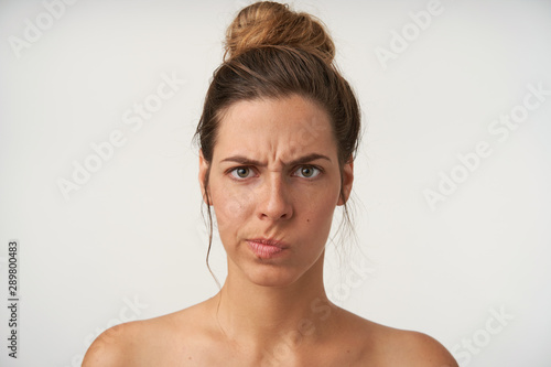 Puzzled attractive young woman with high bun hairstyle standing over white background, looking to camera with serious face and frowning