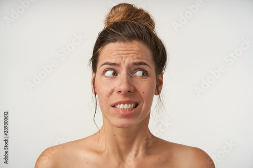 Studio shot of beautiful young woman posing over white background without make-up  looking aside with doubting face  contracting brow and showing teeth