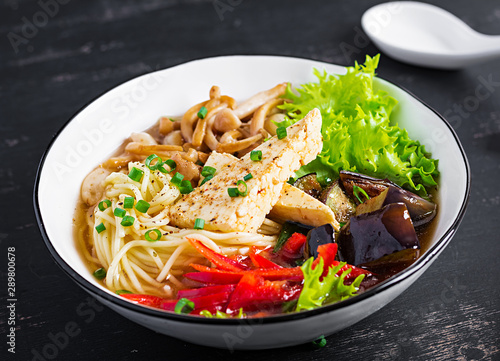 Asian vegan noodles soup with tofu cheese  shimeji mushrooms and fried eggplant in bowl on dark background.