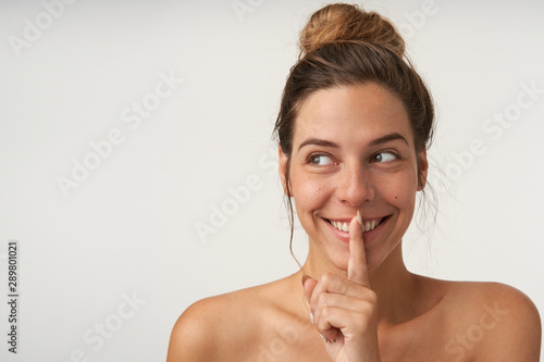 Portrait of young cheerful woman looking aside with broad smile  pretending to keep secret  keeping forefinger near lips  isolated over white background