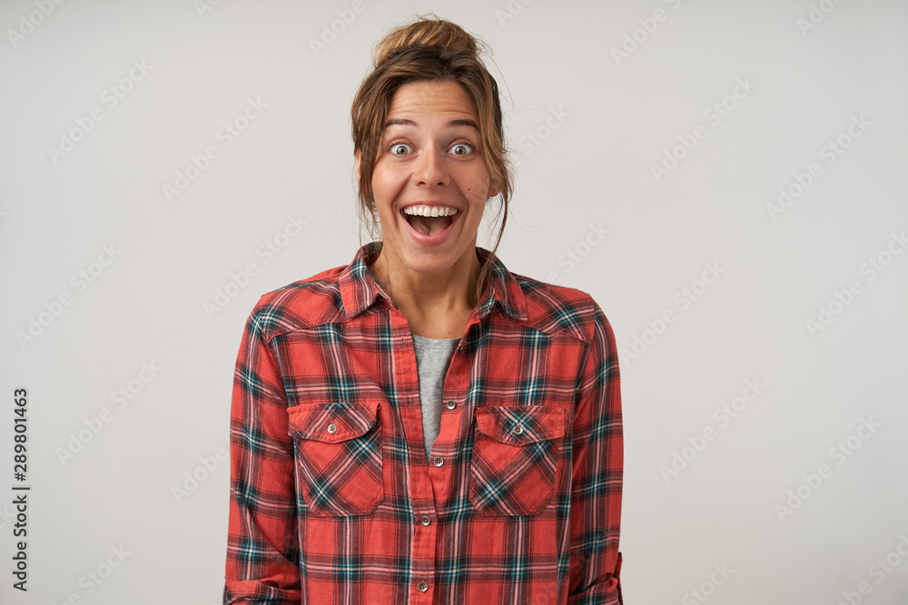 Fototapeta premium Studio shot of surprised pretty young female in checkered shirt, smiling widely to camera with casual hairstyle and natural make-up, isolated over white background