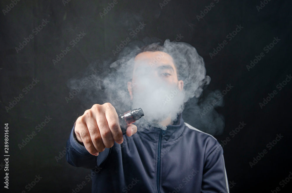 vape and smoke vape electronic e-cigarette or e-cig by a young man with smoke clouds over a black background. vape and smoke vape electronic e-cigarette or e-cig by a young man with smoke clouds over a black background.