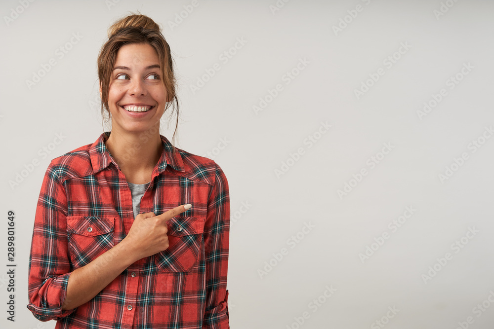 Happy young lovely woman with bun hairstyle posing over white background without make-up, pointing with index finger aside, smiling widely and being in nice mood Happy young lovely woman with bun hairstyle posing over white background without make-up, pointing with index finger aside, smiling widely and being in nice mood