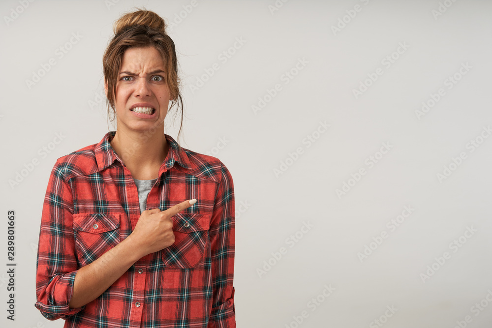 Fototapeta premium Indoor shot of young female showing with forefinger aside, wearing checkered shirt and natural make-up, pouting and frowning, showing disgust on her face