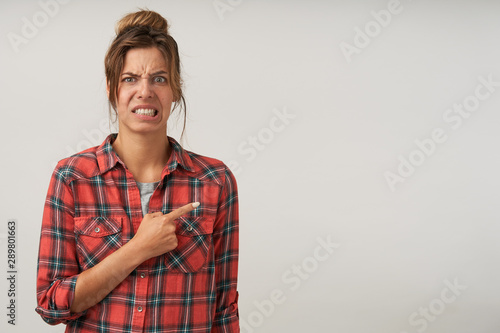 Indoor shot of young female showing with forefinger aside, wearing checkered shirt and natural make-up, pouting and frowning, showing disgust on her face photo