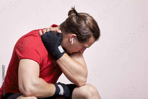 Young caucasian man with white earphones, pony tail, sportswear, boxing bandages, sits in depressed pose. Despair moment, sorrow, hopelessness, despond before or after match. Indoors, copy space.
