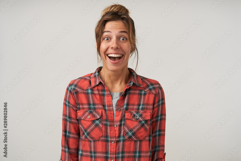 Happy young woman in checkered shirt standing over white background with hands down, looking joyfully to camera with wide mouth opened, having casual hairstyle and natural make-up Happy young woman in checkered shirt standing over white background with hands down, looking joyfully to camera with wide mouth opened, having casual hairstyle and natural make-up
