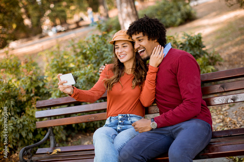 Multiratial loving couple sitting on bench and taking selfie with mobile phone in the autumn city park