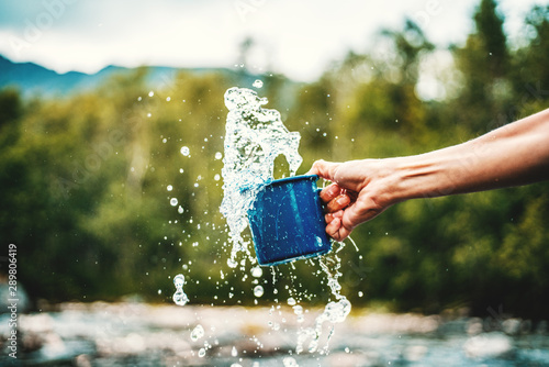 Blue camping mug with clear water, splashes on a forest background. Clear water from a mountain river, ecology concept