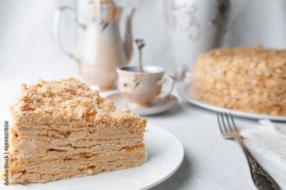 Slice of multilayer Napoleon cake with butter cream. On a white plate. Close-up. Next to a plate is a napkin and a fork. In the background is a cup, teapot and a vase of flowers. Blurred background.