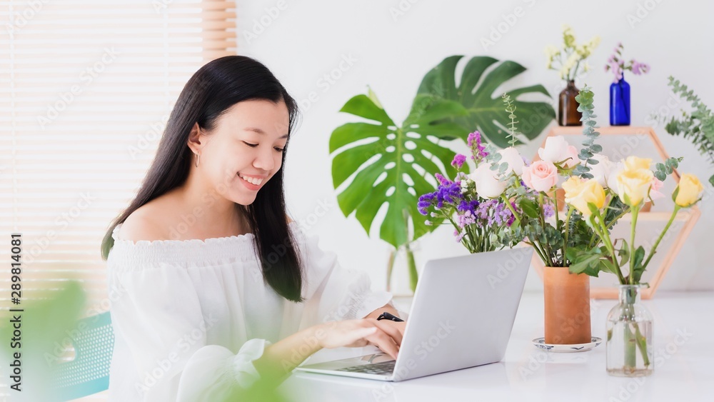 Asian beautiful woman working and using laptop with bouquet rose flowers on white table decoration at home background with smiling face.Lifestyle work to relex at home concept.