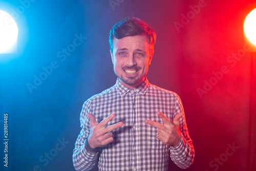 Charming young smiling hipster man is posing against the background of red and blue studio lighting. Concept of pensive man.