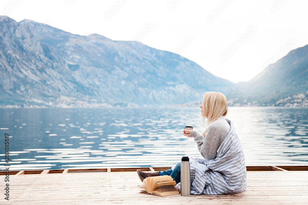 Fototapeta premium Woman on wooden pier by winter sea, mountains. Cozy picnic with coffee, hot beverages, tea or cocoa in thermos and mug, warm plaid, opened book. Girl is enjoying nature, relaxation, reading on beach