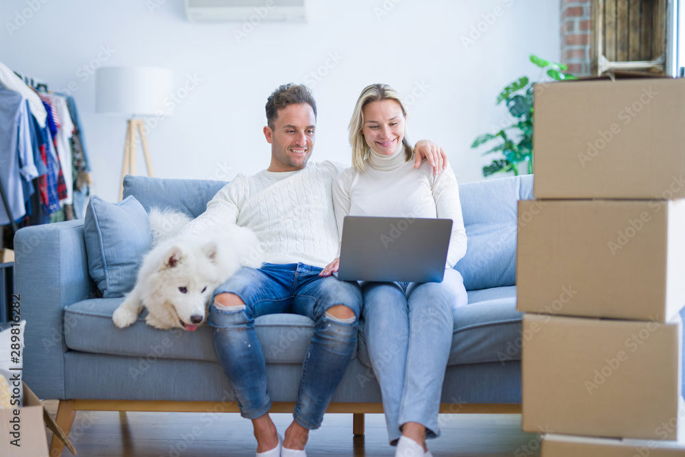Fototapeta premium Young beautiful couple with dog sitting on the sofa using laptop at new home around cardboard boxes
