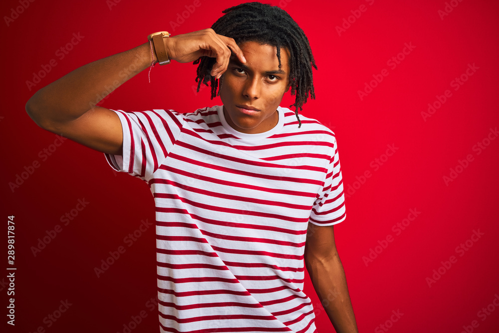 Young afro man with dreadlocks wearing striped t-shirt standing over isolated red background pointing unhappy to pimple on forehead, ugly infection of blackhead. Acne and skin problem Young afro man with dreadlocks wearing striped t-shirt standing over isolated red background pointing unhappy to pimple on forehead, ugly infection of blackhead. Acne and skin problem