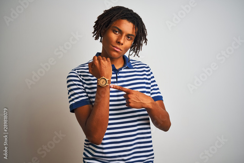 Afro man with dreadlocks wearing striped blue polo standing over isolated white background In hurry pointing to watch time, impatience, looking at the camera with relaxed expression