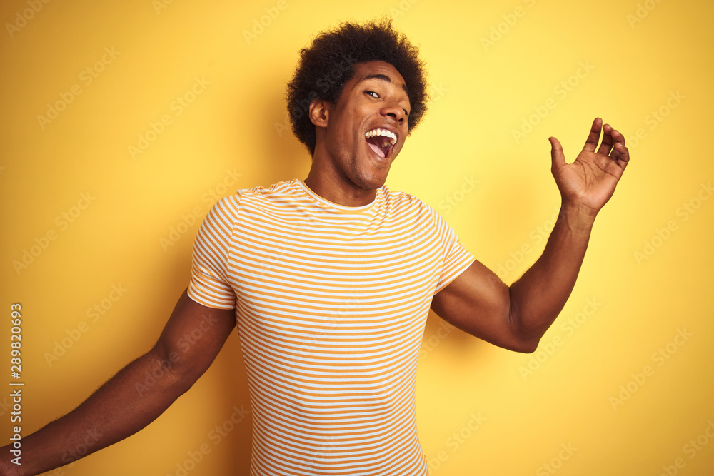 American man with afro hair wearing striped t-shirt standing over isolated yellow background Dancing happy and cheerful, smiling moving casual and confident listening to music American man with afro hair wearing striped t-shirt standing over isolated yellow background Dancing happy and cheerful, smiling moving casual and confident listening to music