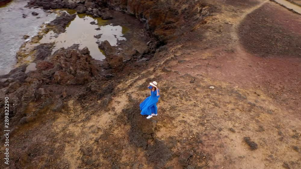 Aerial view of woman in a beautiful blue dress and hat stands on top of a mountain in a conservation area on the shores of the Atlantic Ocean. Tenerife, Canary Islands, Spain Aerial view of woman in a beautiful blue dress and hat stands on top of a mountain in a conservation area on the shores of the Atlantic Ocean. Tenerife, Canary Islands, Spain