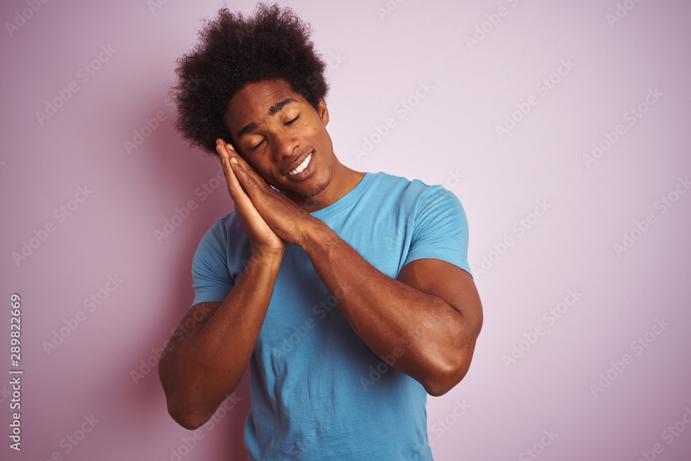 African american man with afro hair wearing blue t-shirt standing over isolated pink background sleeping tired dreaming and posing with hands together while smiling with closed eyes. African american man with afro hair wearing blue t-shirt standing over isolated pink background sleeping tired dreaming and posing with hands together while smiling with closed eyes.