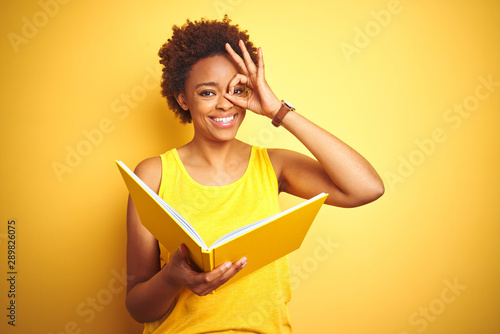 African american woman reading a book over yellow isolated background with happy face smiling doing ok sign with hand on eye looking through fingers