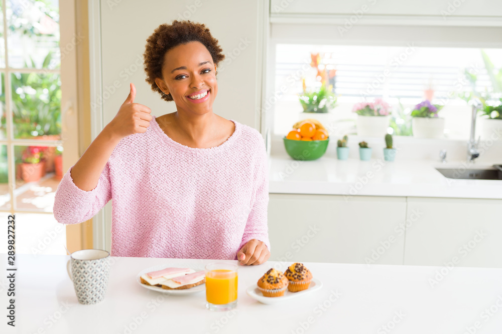 Young african american woman eating breaksfast in the morning at home doing happy thumbs up gesture with hand. Approving expression looking at the camera with showing success.