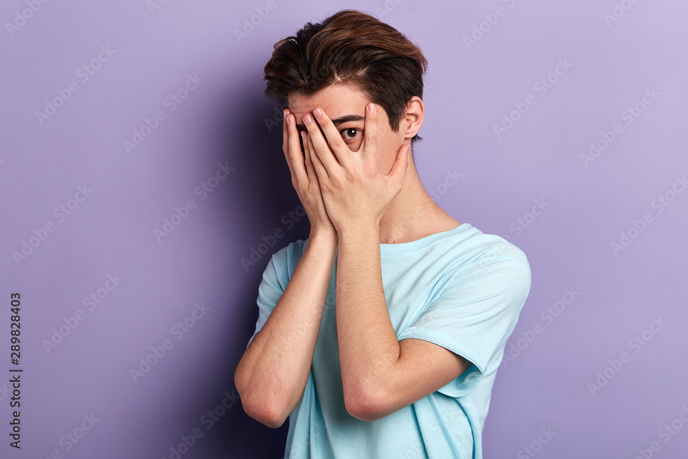 Handsome manhides face with hands,lookig through palms. peeks through fingers, wears fashionable T-shirt, feels shy, isolated over blue background.fear, fobia concept. isolatedd blue background Handsome manhides face with hands,lookig through palms. peeks through fingers, wears fashionable T-shirt, feels shy, isolated over blue background.fear, fobia concept. isolatedd blue background
