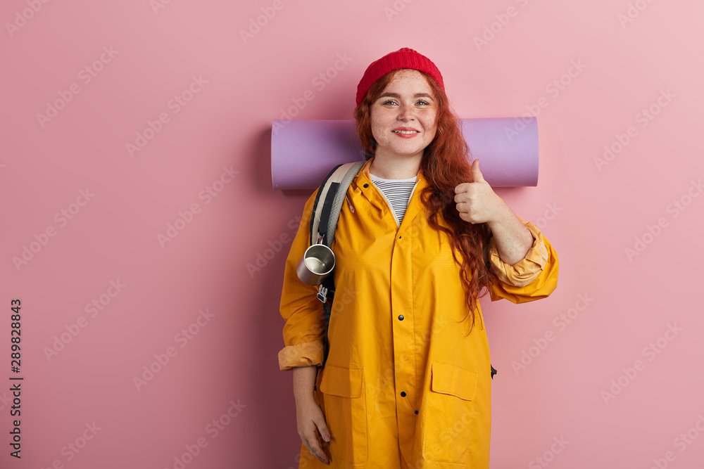 cheerful ginger woman wearing mat a, backback, yellow jacket, cap, shirt hiker shows thumb up, tourits likes her holiday,adventurer expresses positive feeling and emotion. isolated pink background. cheerful ginger woman wearing mat a, backback, yellow jacket, cap, shirt hiker shows thumb up, tourits likes her holiday,adventurer expresses positive feeling and emotion. isolated pink background.