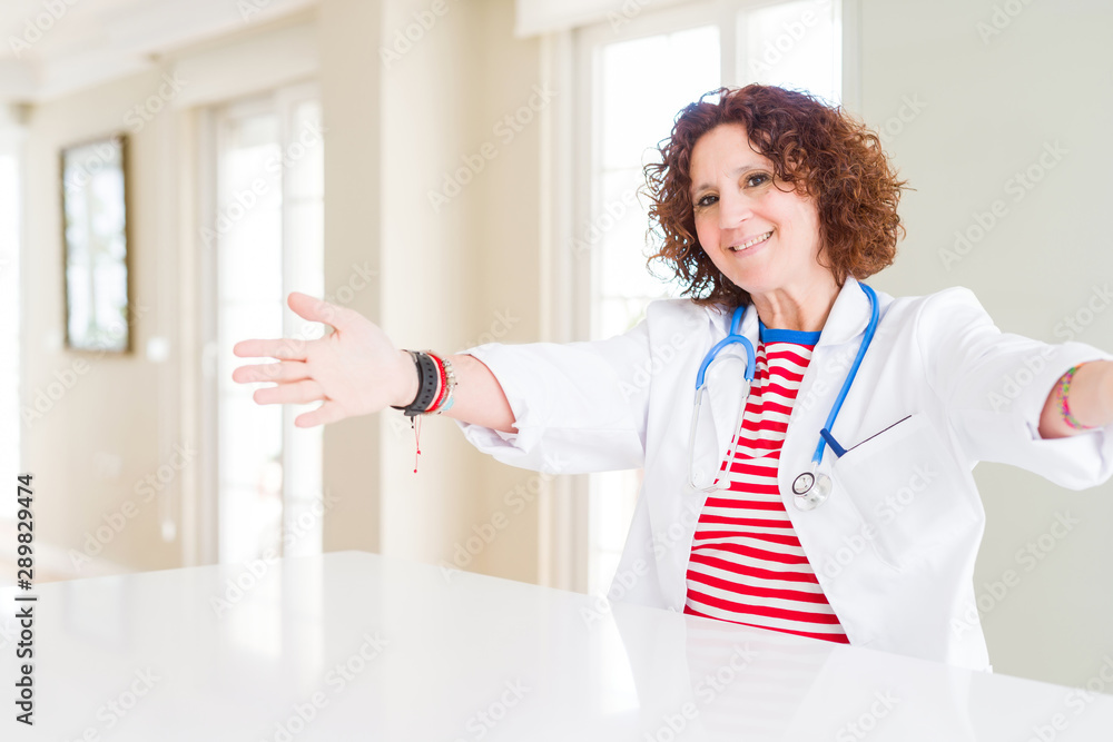 Senior doctor woman wearing medical robe at the clinic looking at the camera smiling with open arms for hug. Cheerful expression embracing happiness. Senior doctor woman wearing medical robe at the clinic looking at the camera smiling with open arms for hug. Cheerful expression embracing happiness.