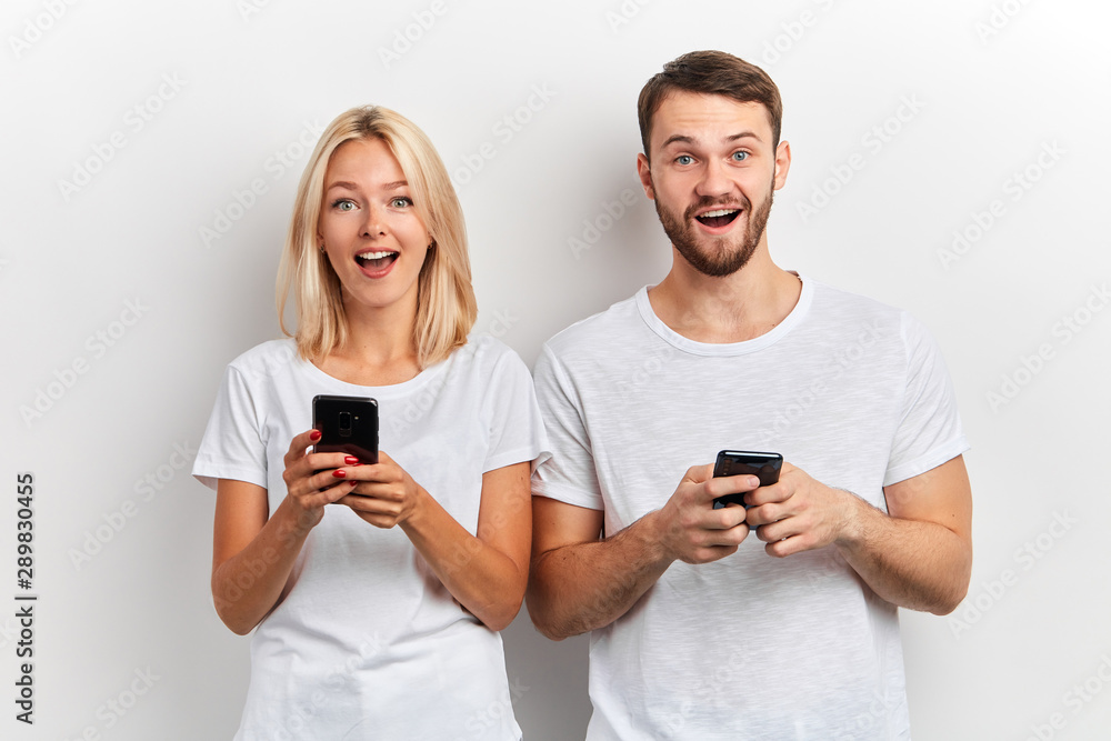 Photo of beautiful excited happy man and woman with wide open mouth smiling while both using smart phones isolated over white background. happiness, positive feeling and emotion, sale discount Photo of beautiful excited happy man and woman with wide open mouth smiling while both using smart phones isolated over white background. happiness, positive feeling and emotion, sale discount