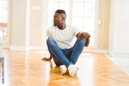 Handsome african american man sitting on the floor at home looking to side, relax profile pose with natural face with confident smile. © Krakenimages.com