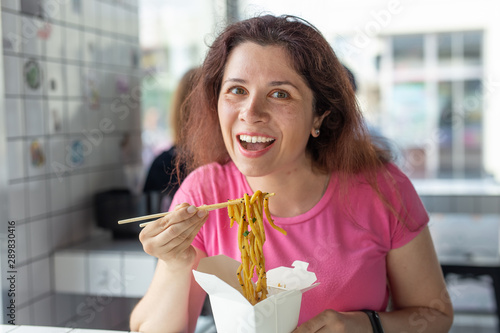 Portrait of a young cheerful girl eating chinese noodles in a cafe and looking out the window. The concept of healthy Asian cuisine.